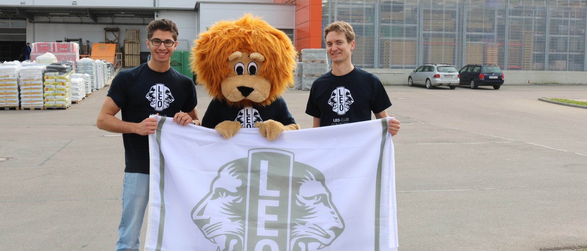 Zwei Leos mit dem Löwen Maskottchen in der Mitte halten ein Banner mit Leo Logo